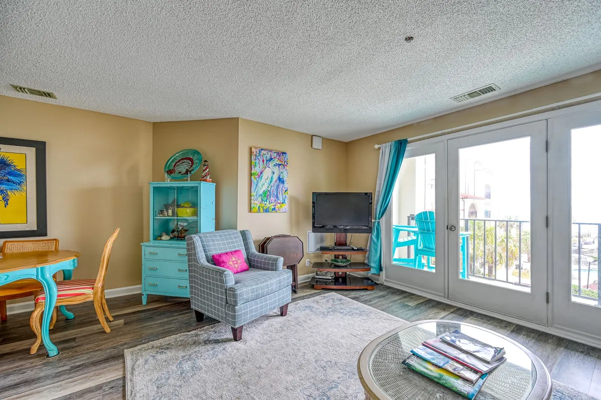 A cheerful, sunlit room with large windows letting in sunrise views, colorful artwork, and turquoise furniture. The openness and brightness of the space suggest a seamless connection to the outdoors, hinting at morning coffee before heading out for beach activities.