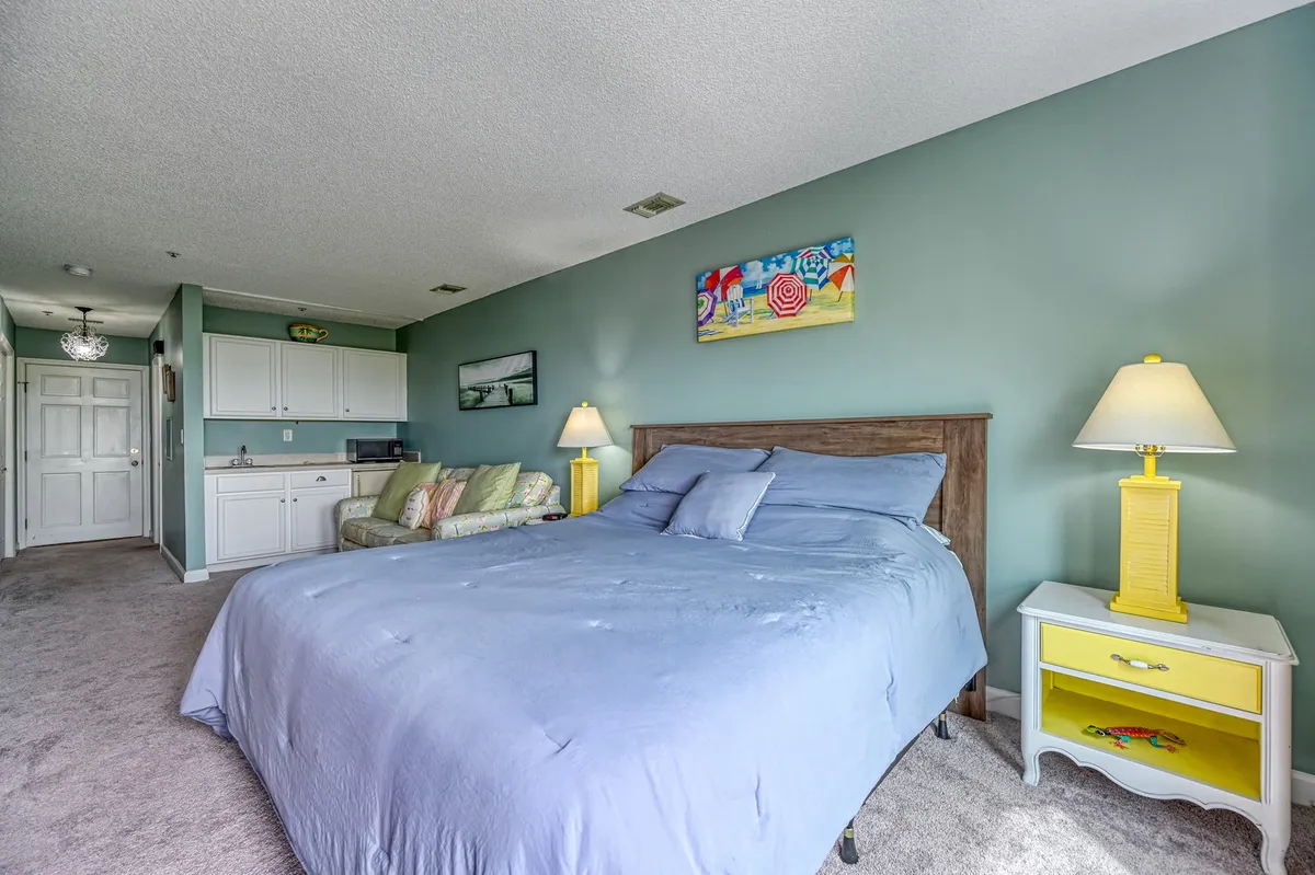 A spacious primary suite featuring a king-sized bed with plush bedding, a yellow bedside lamp adding warmth, vibrant wall art above the headboard, and a compact kitchenette equipped with a sink and coffee maker in the corner. Sunlight filters through the window, creating a bright and inviting atmosphere ideal for relaxation and family comfort.