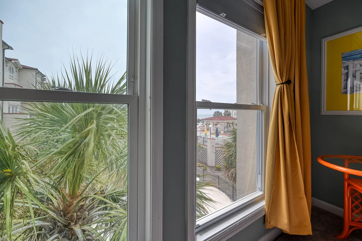 This image features a bright, cheerful space with floor-to-ceiling windows that frame a palm tree and glimpses of the outdoors. The scene is set for a relaxing morning with a cup of coffee at an orange circular table, surrounded by vibrant yellow curtains that fill the space with light. The connection to the outside—through the palm tree and view beyond—suggests a strong link between the cozy interior and the tranquil coastal setting just outside.