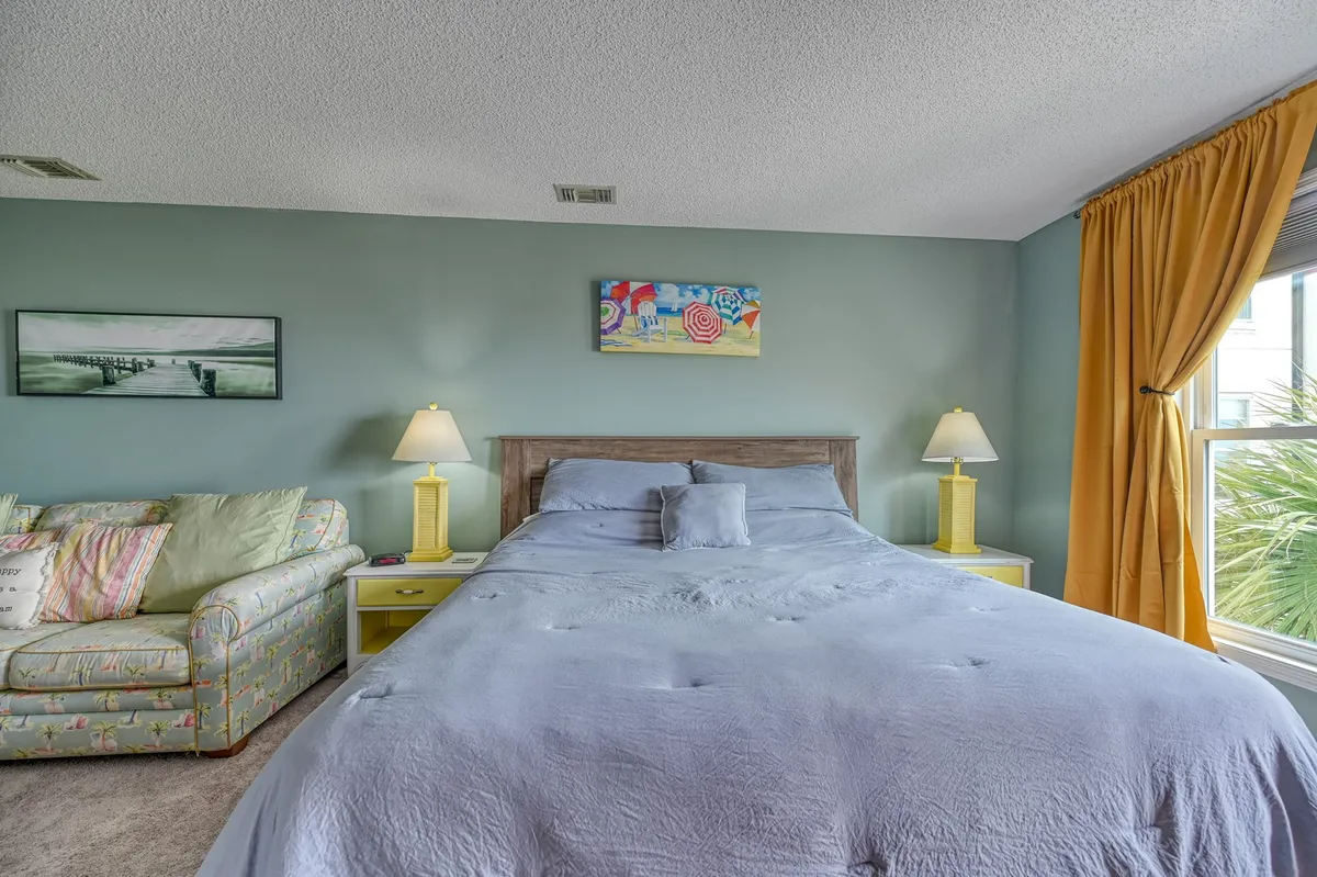 A cozy guest bedroom with a queen bed, soft lamps, and light-diffusing curtains. The room is styled with inviting bedding, a small sofa for extra comfort, and framed artwork that adds a personal touch. Warm natural light streams in, creating a restful atmosphere perfect for guests or families.