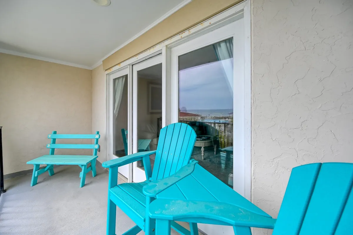 An exterior balcony scene with turquoise chairs facing a scenic view beyond the sliding glass doors. The tranquil setup suggests a place for family members to relax, socialize, and enjoy the ocean breeze after active outdoor play.