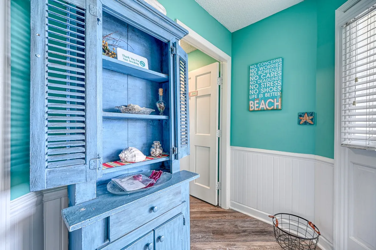 A bright entryway with a blue wooden cabinet, beach-inspired decor, and motivational wall art sets a welcoming, coastal mood. The area is ideal for storing beach gear and evokes a sense of anticipation for days spent exploring the shore.