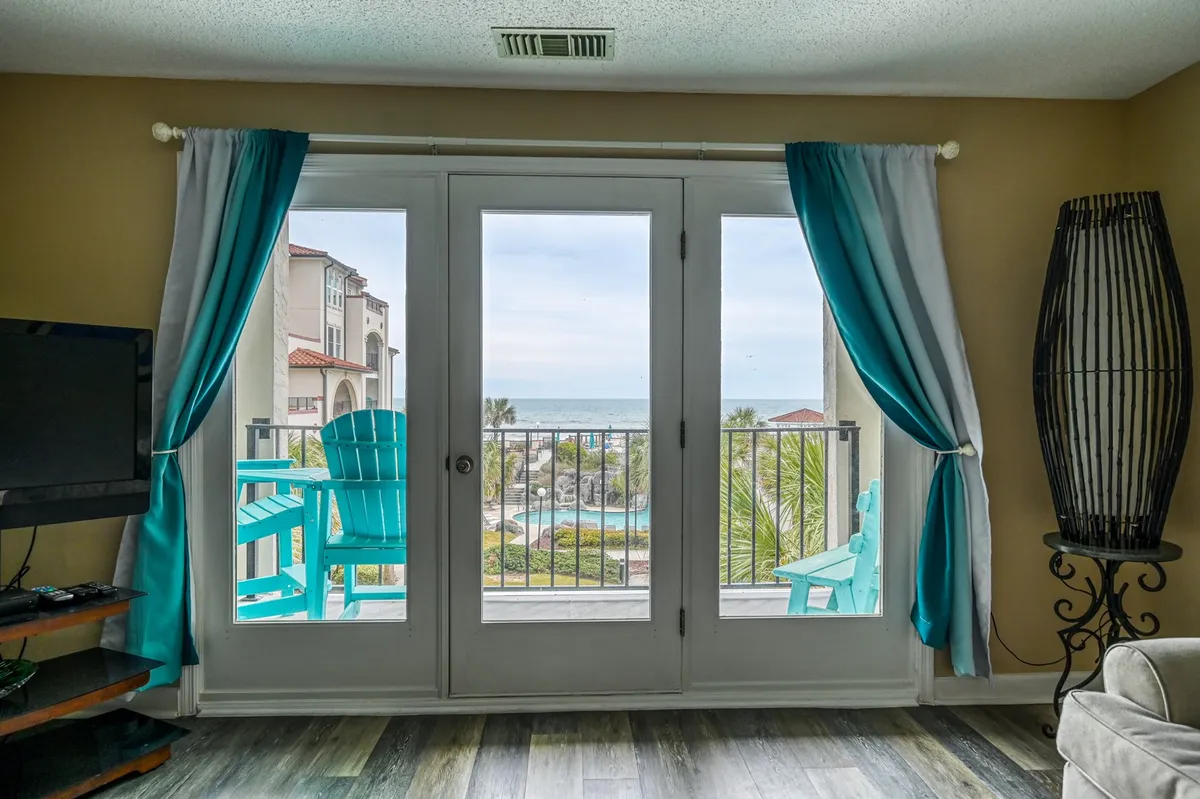 This image features double doors opening to a balcony with blue chairs, offering an inviting place to relax and enjoy the ocean views. The interior space is calm and inviting, with a television and decorative lamp creating a welcoming environment for gathering or downtime.