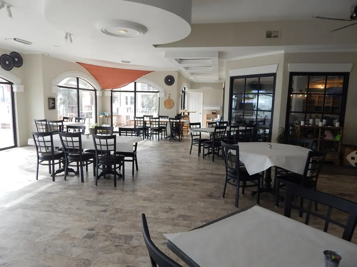 A bright, stylish dining area with tables draped in white tablecloths, flooded with natural light from large windows. Subtle decorative touches hint at an upscale yet inviting environment, ideal for social gatherings or enjoying a meal.