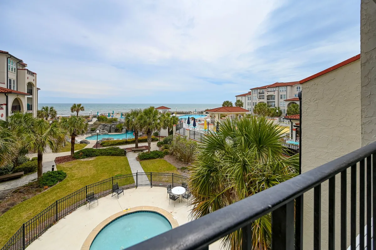 A relaxed view from a condo balcony overlooking the swimming pool and landscaped gardens below, with the ocean stretching into the distance. The scene is inviting, showcasing the transition from private living to shared vacation amenities.