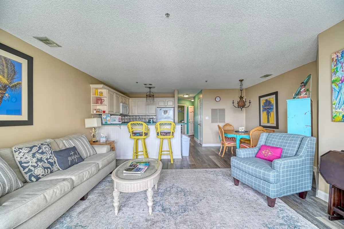 This image showcases the main living area with a sofa adorned in decorative pillows, a blue accent chair, yellow bar stools, a dining table set for group meals, and vibrant artwork on the wall. The space is open and inviting, ideal for both relaxing and socializing.
