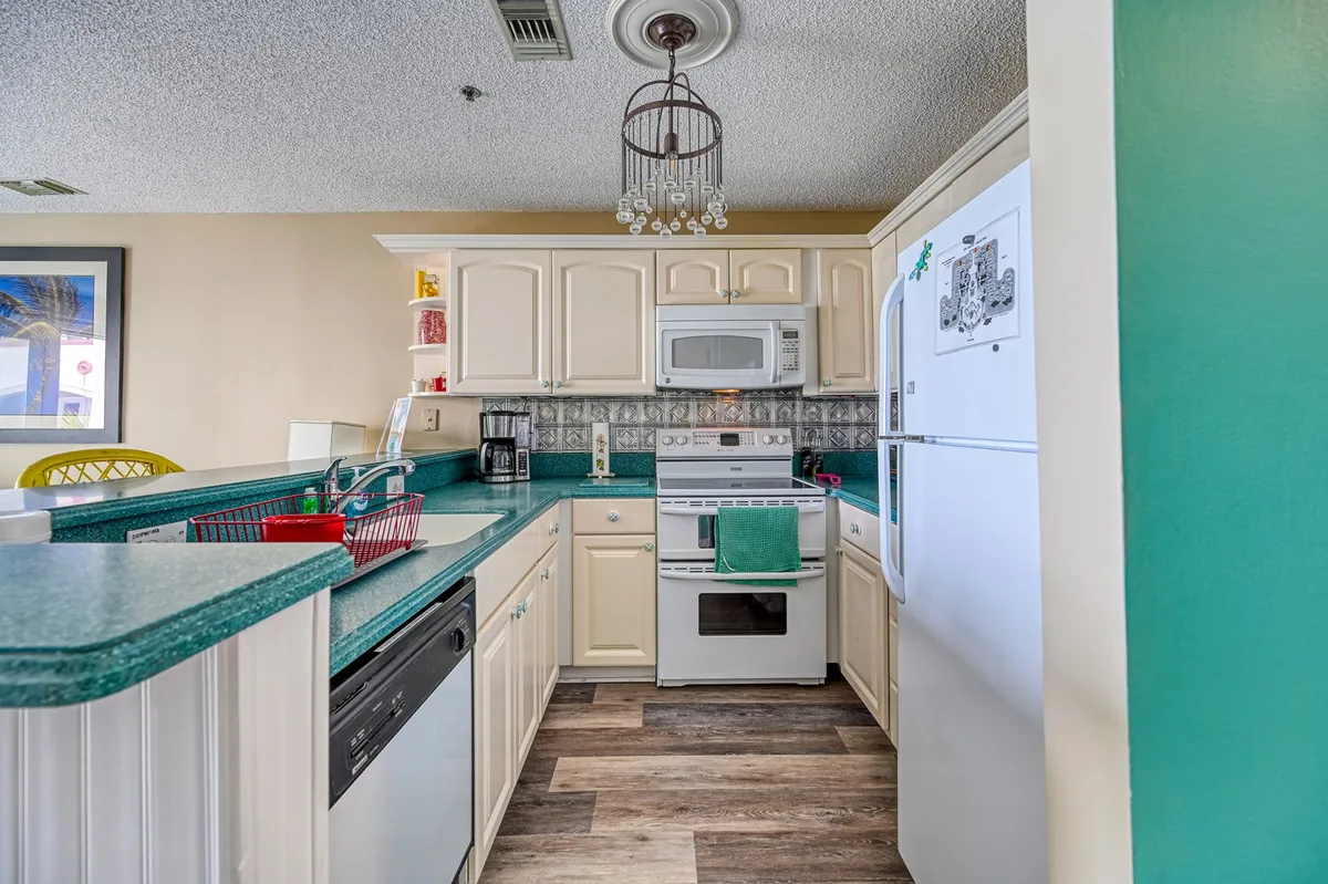 The kitchen is the focal point here, featuring a teal countertop island, white appliances, colorful wall décor, and a lively chandelier. The inviting setup is perfect for cooking, entertaining, and shared family meals.