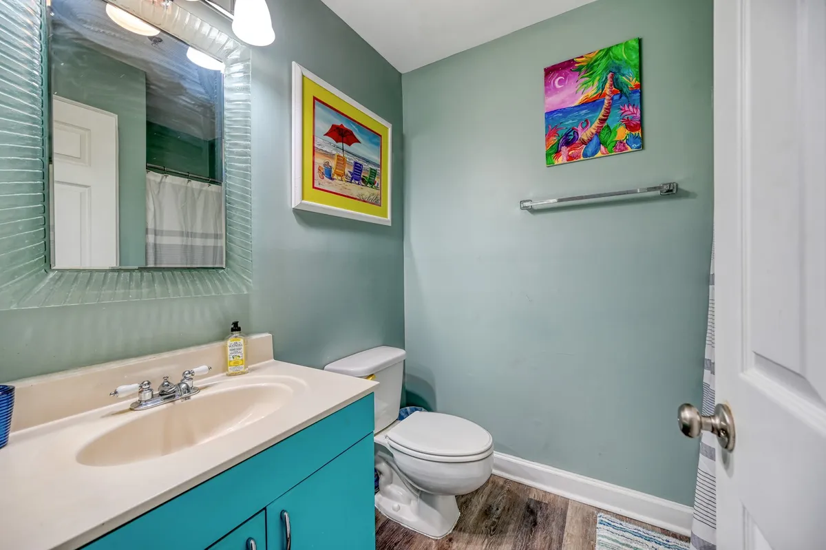 A bright, inviting bathroom featuring a bold turquoise vanity, a large mirror, and colorful wall art. The space feels clean and fresh, with ample counter space for shared use and a calming atmosphere perfect for morning or post-beach routines.